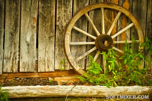 3353114-antique-wagon-wheel-leaning-against-an-antique-wooden-barn-with-weeds-growing-in-front