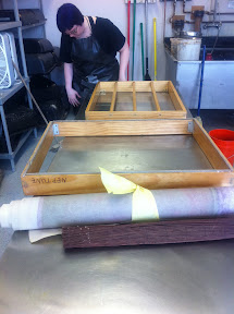 This is me setting up the mould (or mold) and deckle to make paper on.  The mould is a screen that the pulp fibers can sit on while the water drains out.  The deckle is a frame that goes over the mould so that all the pulp fiber is held in place.  Generally, you dip the mould and deckle into a vat of pulp, but here, we are using a "deckle box" set up, where we pour the pulp into the mould instead.