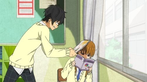 Haru stands over Shizuku who is sitting at her desk intently reading a book and ignoring him while he hits her on the head with a school book