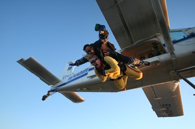image shows two people jumpsing out of a plane, the guy behind delighted, the guy in front of him, dressed in yellow, scared out of his tree, like I'll be doing