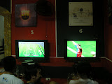 Este es un bar llamado PS3. Esta lleno de consolas y la gente juega mayoritariamente al futbol, y con los equipos espanyoles. En el monitor 6, el Barca acaba de marcar. Encima, un poster del Alicante o el Valencia, y a la izquierda, mas Barca.