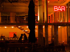 [ROT BELEUTETE BAR MIT REINER GLASSFASSADE. IN DER BAR SITZT EIN PAAR UND KÜßT SICH.]