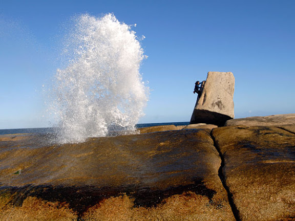 A woman climbing a boulder with a wave exploding Bicheno Tasmania