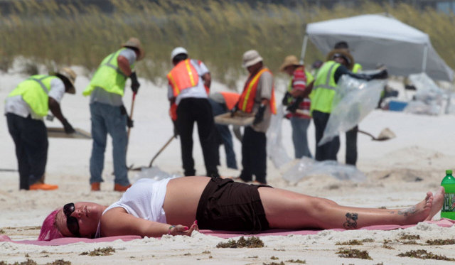 Frances Sexton of Indianapolis, Ind., lies on the beach as oil cleanup workers clean the beach in the background in Pensacola Beach, Fla., Wednesday, July 7, 2010. Oil washed ashore overnight leaving an ugly stain that brought out hundreds of BP workers to clean. AP Photo / Dave Martin