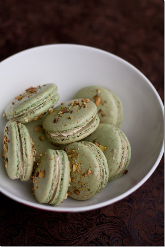 A white bowl full of Pistachio macarons topped with pistachios. 