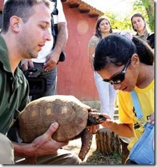 Homem segura uma tartaruga; moça cega passa a mão no animal