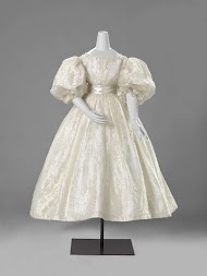 Trouwjapon van wit zijden gaas met een ingeweven wit satijnen bloemen- en randpatroon, met twee om elkaar slingerende bloem- en bladranken, vanuit golvende S-voluten