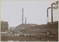 Exterieur van fabrieksgebouwen en rokende schoorstenen, met op de voorgrond houten vaten