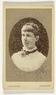 Ovaal studioportret van een jonge vrouw in een witte jurk, om haar nek draagt zij een lint met een bloem