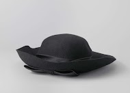 Hoed van zwart vilt met satijnen band om bol, die middenvoor eindigt in een platte strik met bijpassende hoedenspeld