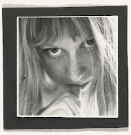 Gezicht van een vrouw met blond haar en grote ogen, haar duim tegen haar lippen