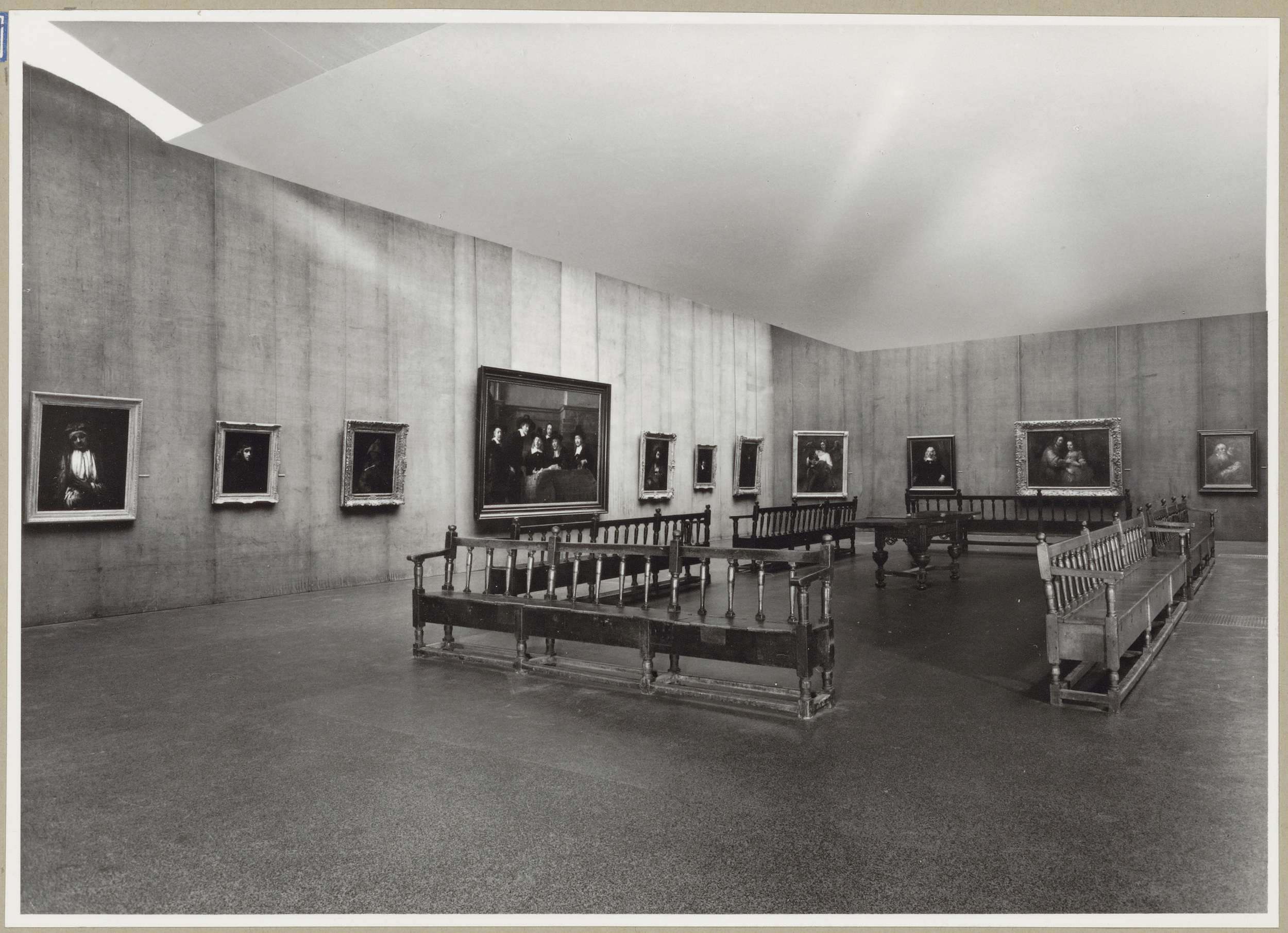 Zaal met elf schilderijen en een tafel te midden van banken