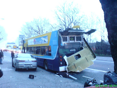 Photo shows roof of bus from rear on the road. Back window is totally smashed as is the surrounding metal. A fire engine can be seen in the background. Photo taken at 08:12