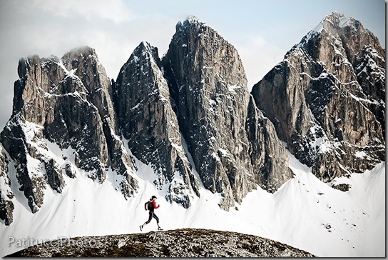 A female trail runner in the Italian Dolomites with the Geislergruppe, covered in snow, in the background.